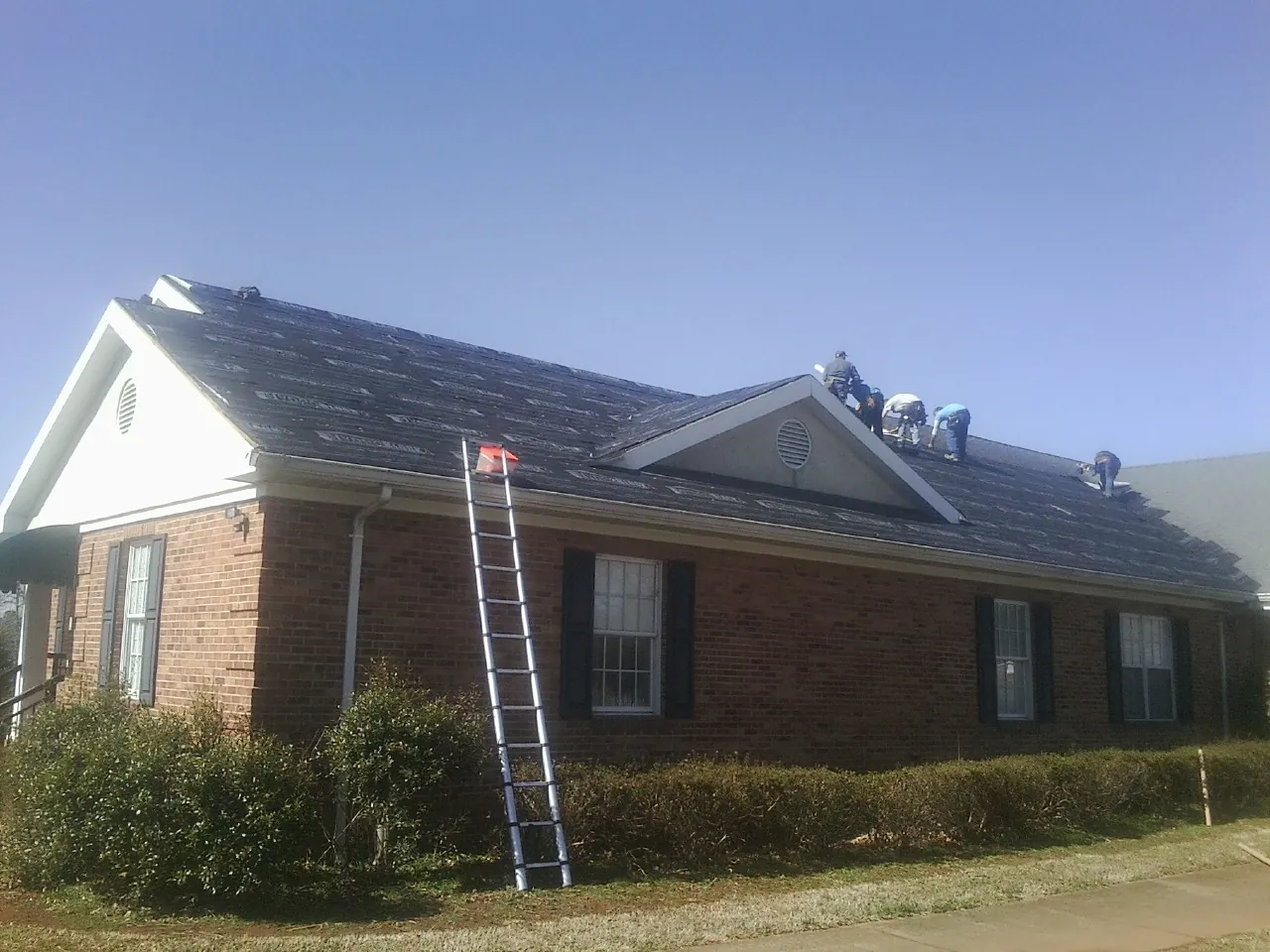 Crew tearing shingles off a brick ranch roof with a ladder set — Emergency Roof Repair work by our crew in Larchwood