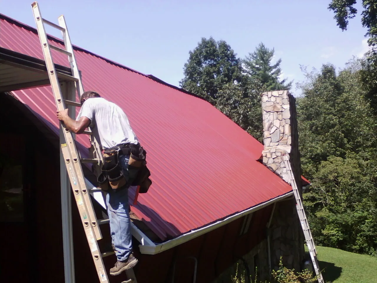Roofer on a ladder beside a red metal roof and stone chimney — Soffit & Fascia Repair by Roofing Brothers in Larchwood